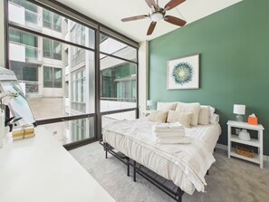 A bedroom with a large window, a bed with white linens, a ceiling fan, and a framed artwork on the wall.