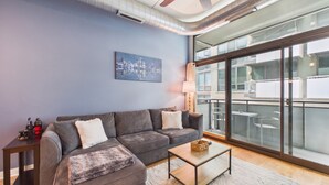 Un salon moderne avec un canapé gris, une table basse, un tableau et une grande fenêtre donnant sur un espace de bureaux.