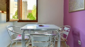 Une pièce avec une table et des chaises, une fenêtre dont le volet est ouvert, et un mur violet orné d’un tableau encadré.