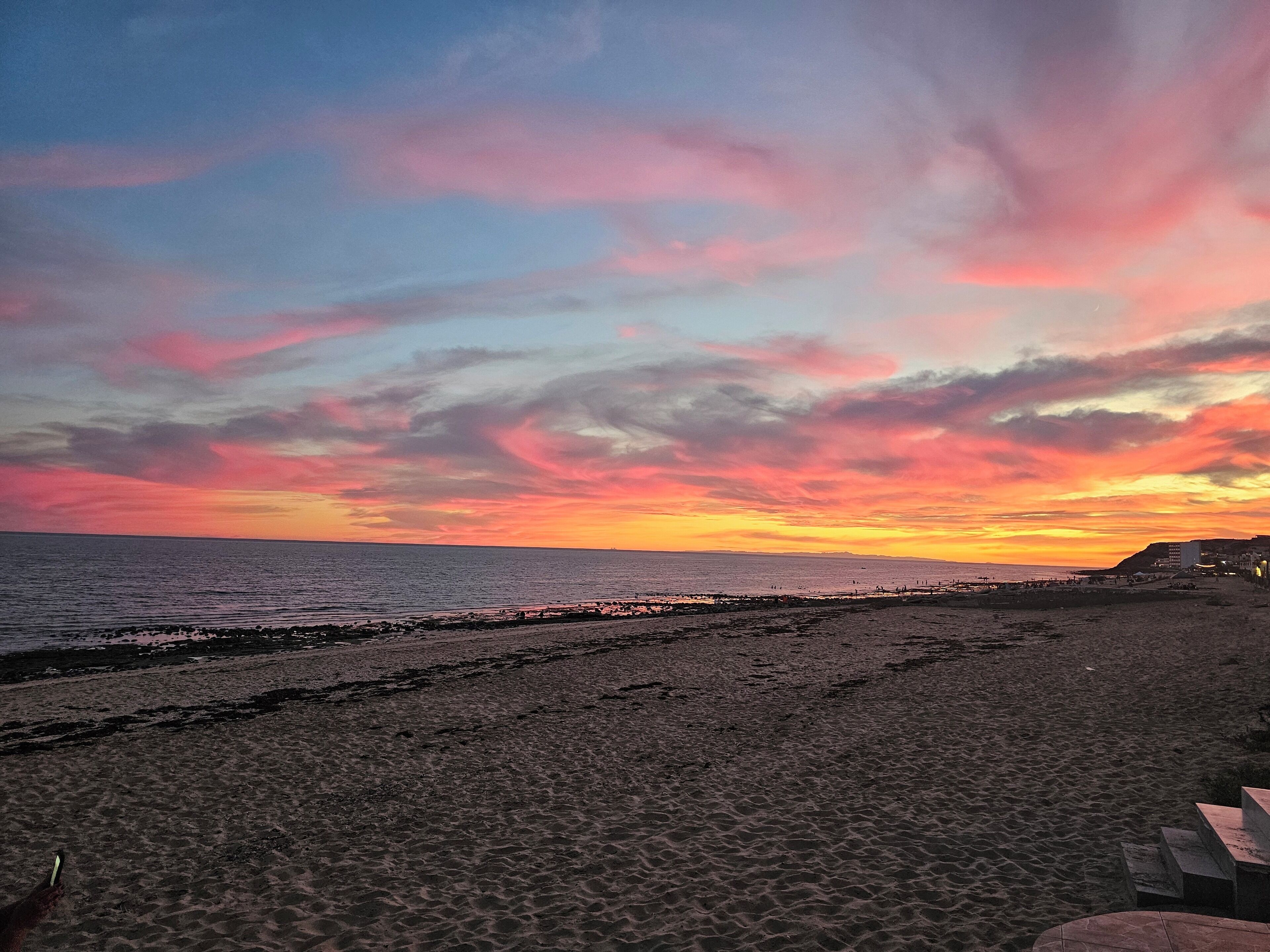Una playa con atardecer, mar tranquilo y edificios a lo lejos.