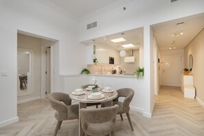 A dining area with a round table set for four, surrounded by four chairs. A kitchen is visible in the background, and a hallway leads to a door. - Stayshort - Refined 1 Bedroom Living in Sports City That Sleeps 4 (Dubai)