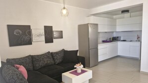 A modern living room with a black sectional sofa, a white coffee table, and a kitchen area with white cabinets and a stainless steel refrigerator. - Apartment Slanica Beach Escape - One Bedroom Apartment With Terrace And Sea View (Murter)