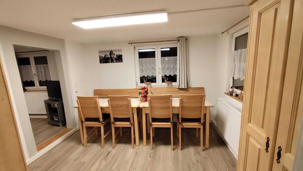 A compact kitchen with a dining area, featuring a wooden table and chairs, a small refrigerator, and a window with curtains. - Vacation home in the heart of the Allgäu in Heimenkich (Heimenkirch)