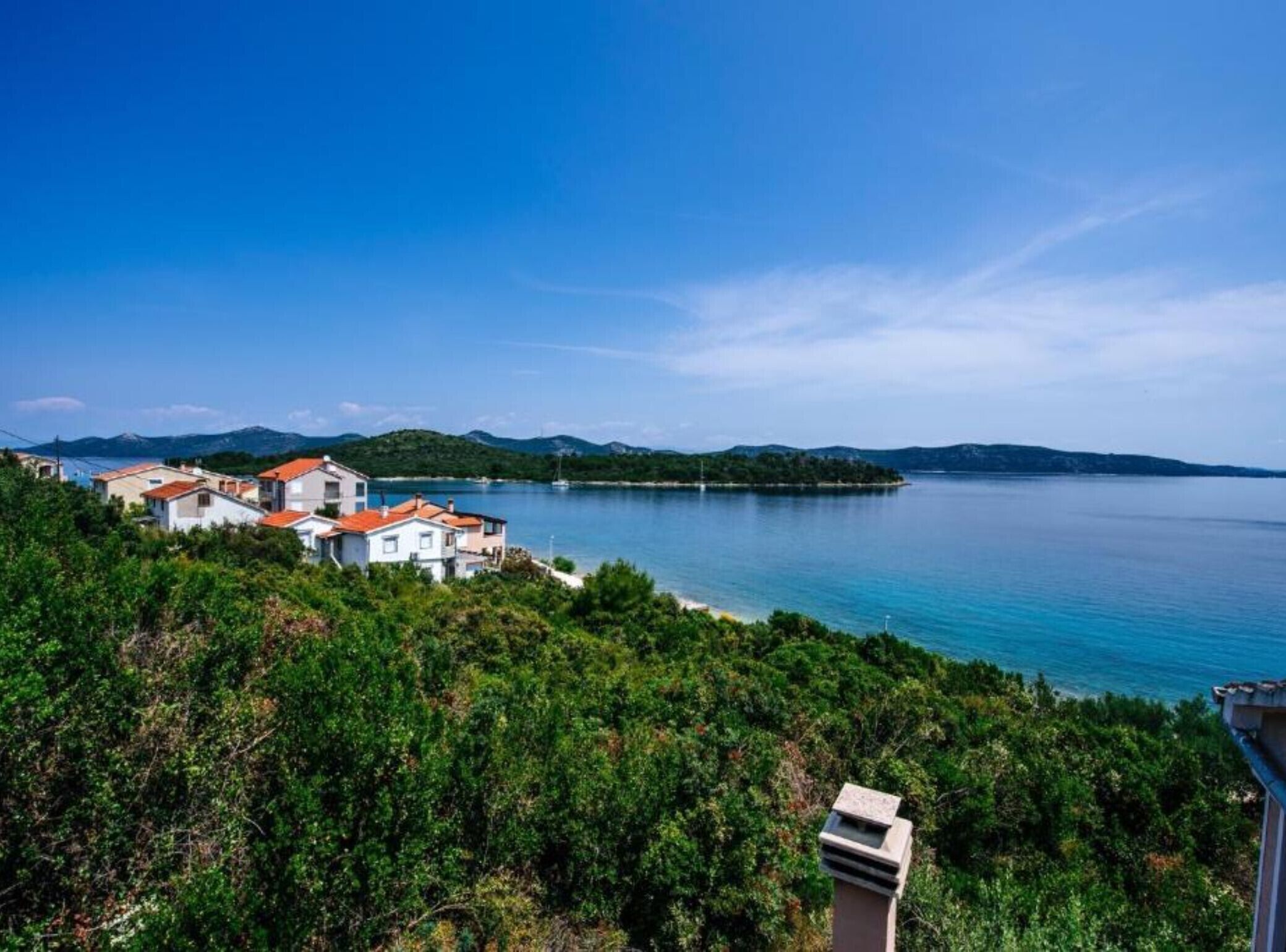 A coastal village with houses, a clear blue sea, and a hilly landscape in the background.