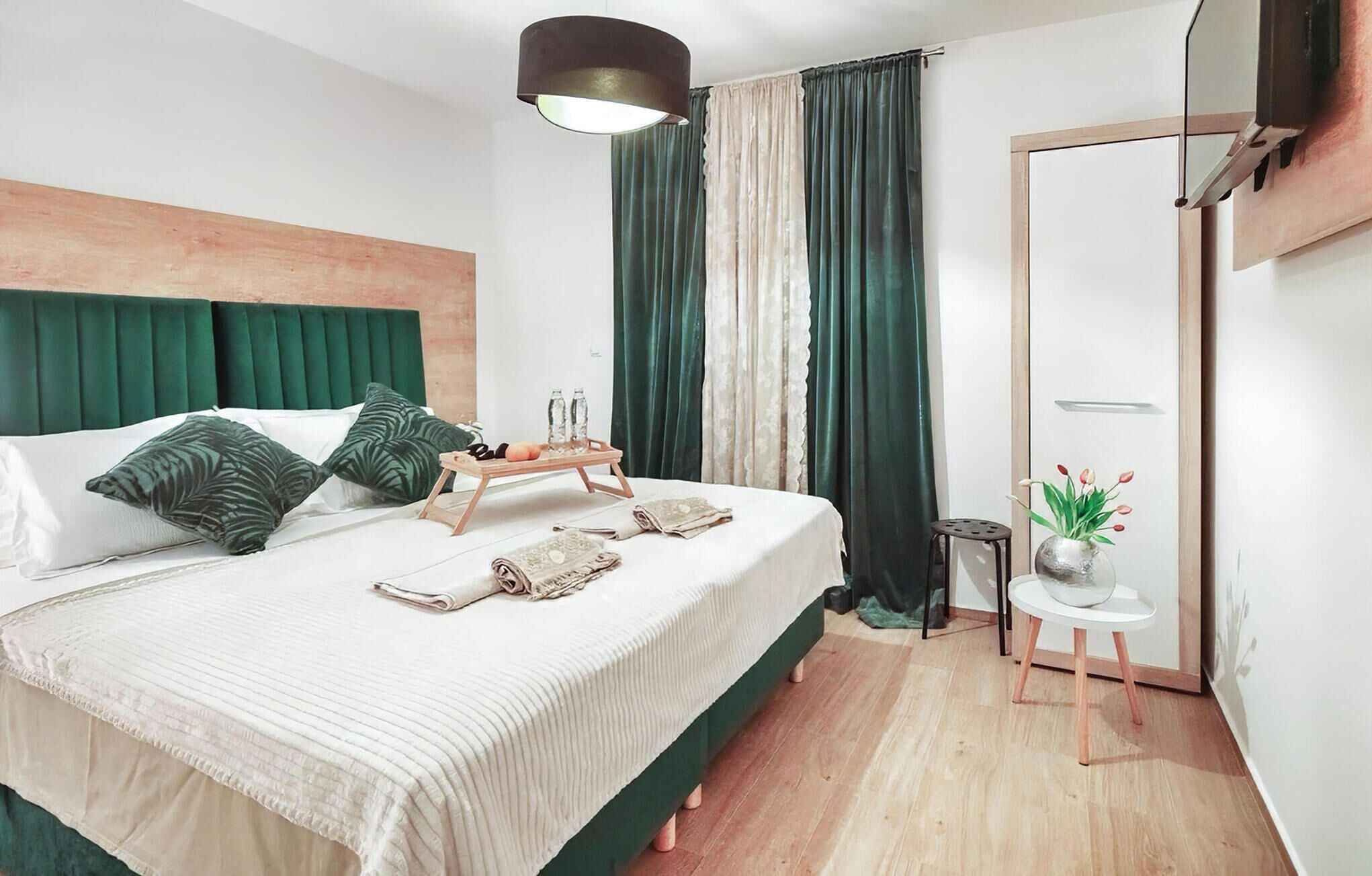 A modern bedroom with a large bed, green curtains, a small table with a tray, and a potted plant.
