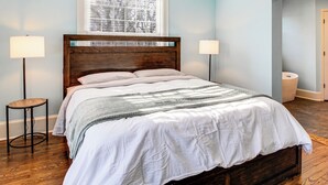 Quarto com cabeceira de madeira, uma janela grande com persianas, mesa de cabeceira e criado-mudo.