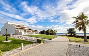 A modern building with a paved pathway leading to a body of water, surrounded by greenery and palm trees. - Lovely apartment in Medulin with house sea view (Medulin)