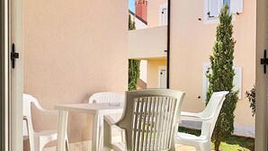Une terrasse avec des chaises et une table en plastique blanc, un mur de couleur beige et une vue sur un immeuble doté d’une cheminée.