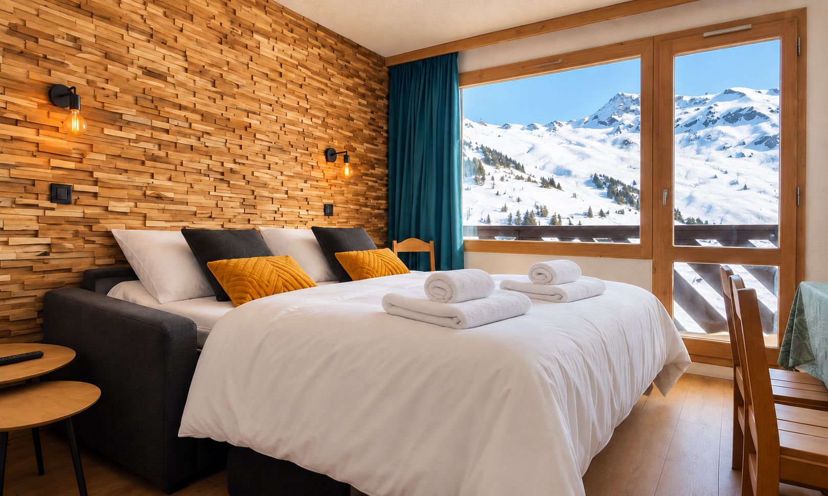 A hotel room with a wooden wall, a bed with white bedding, a sofa with cushions, a small table, and a view of snowy mountains through a large window.