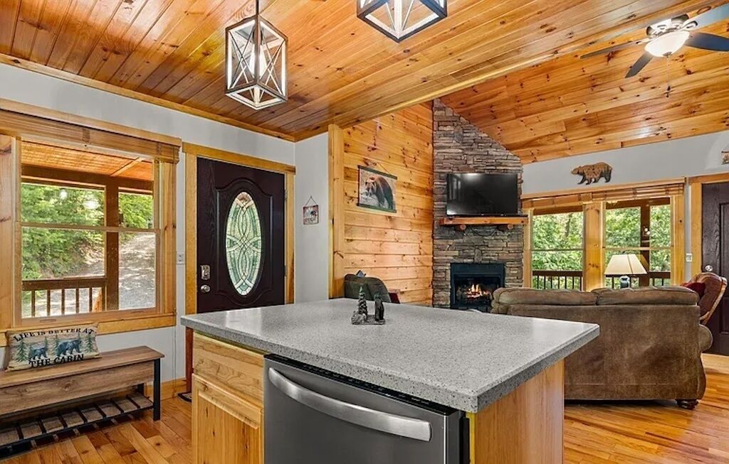 A cabin interior with a kitchen island, stone fireplace, wooden walls, and large windows.