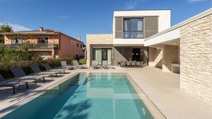 Un espace piscine moderne, doté de chaises longues et d’une piscine à eau claire, entouré de murs en pierre et d’un bâtiment à deux étages avec de grandes fenêtres.