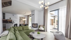 Un salon moderne avec un canapé d’angle, une table basse ronde et un coin salon agrémenté d’un pouf blanc. On aperçoit également un coin repas avec une table et des chaises, ainsi qu’une cuisine au fond.