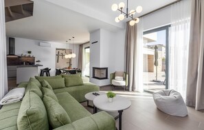 Ein modernes Wohnzimmer mit einer Eckcouch, einem runden Couchtisch und einer Sitzecke mit einem weißen Pouf. Es gibt einen Essbereich mit Tisch und Stühlen, und im Hintergrund eine Küche.