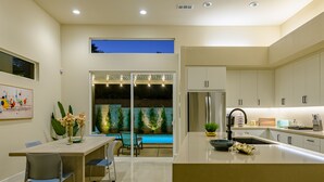 A modern kitchen with a large island, a dining area with chairs, and a view of a swimming pool at dusk. - Brand New! Chic, Gated & Safe. Family & Pet Friendly, Sleeps 9 with Rec. Room… (Indio)