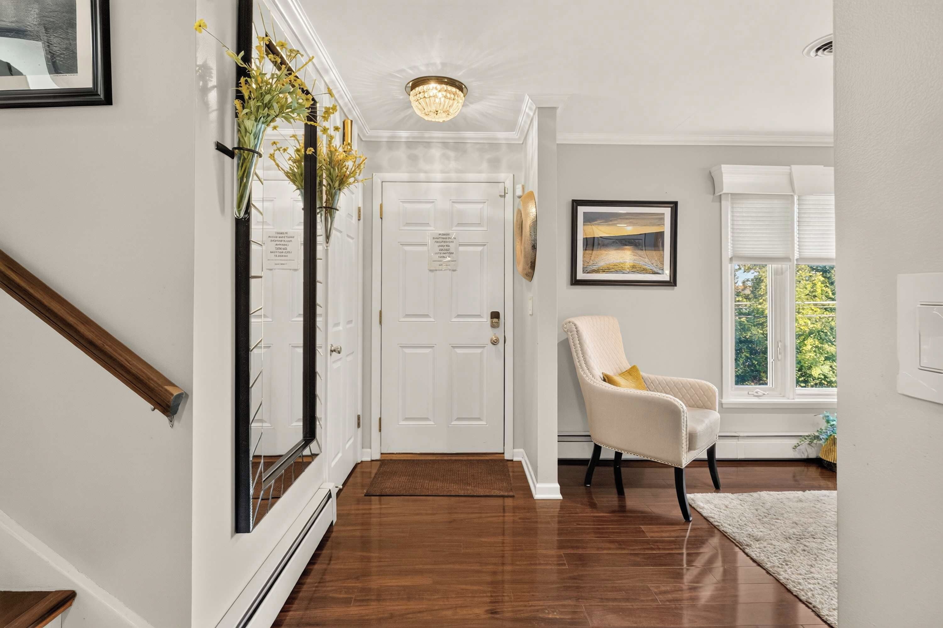 A hallway with a wooden floor, a white door, a white chair, a framed picture on the wall, and a window with blinds.