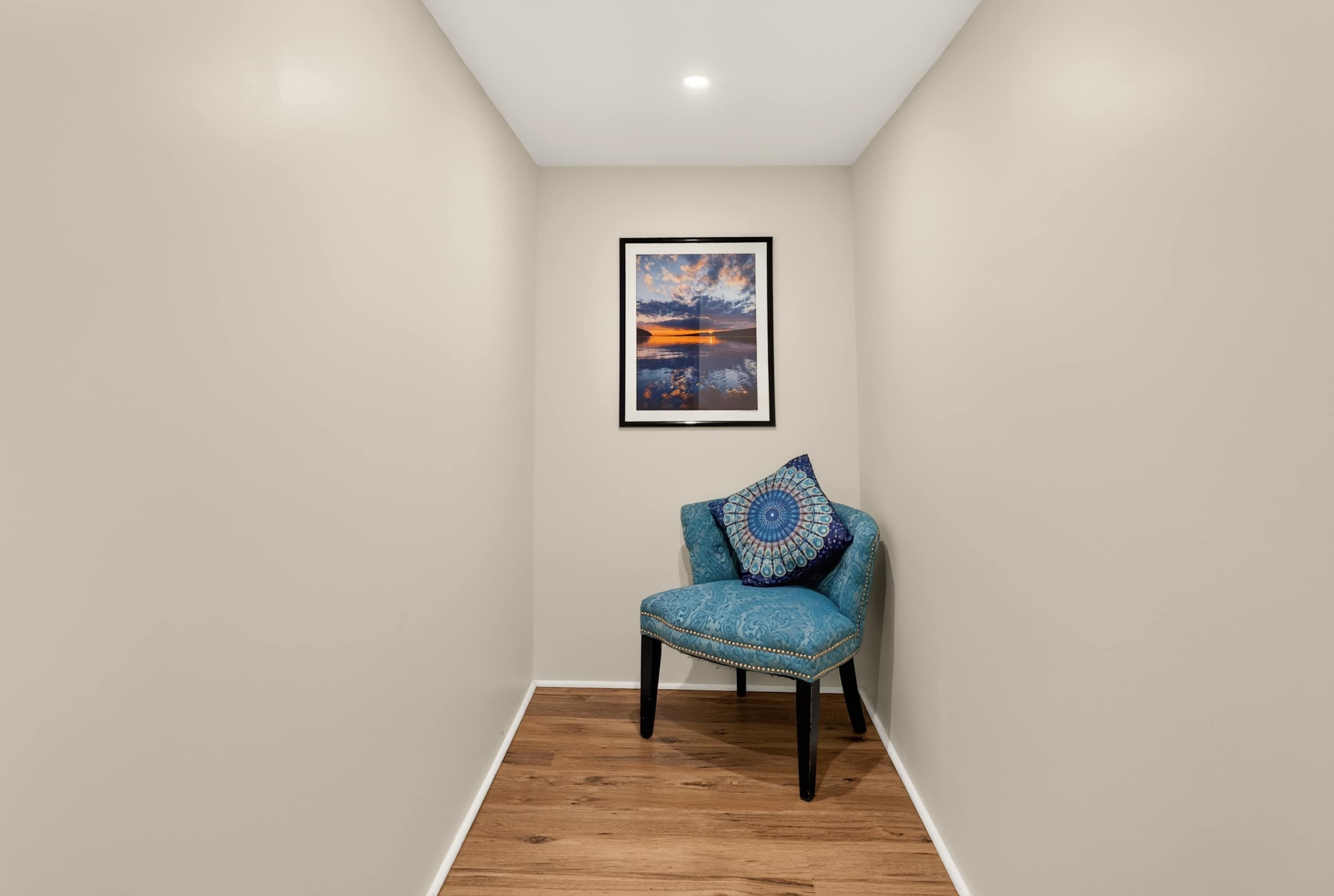 Una habitación con un cuadro enmarcado en la pared, un sillón tapizado azul con un cojín decorativo y suelo de madera.