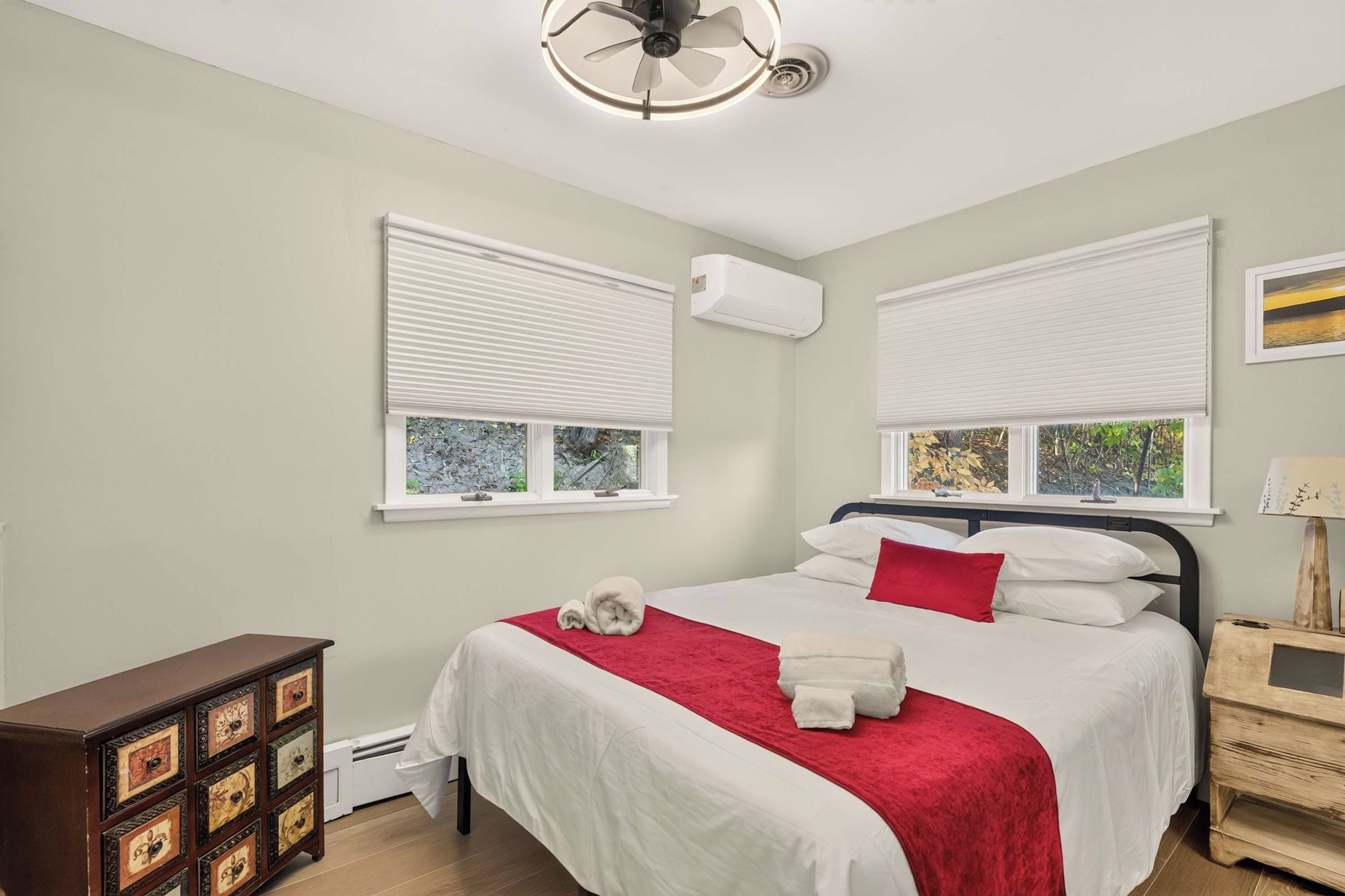 A bedroom with a bed, two windows with blinds, an air conditioning unit, a bedside table with a lamp, and a decorative wooden chest.