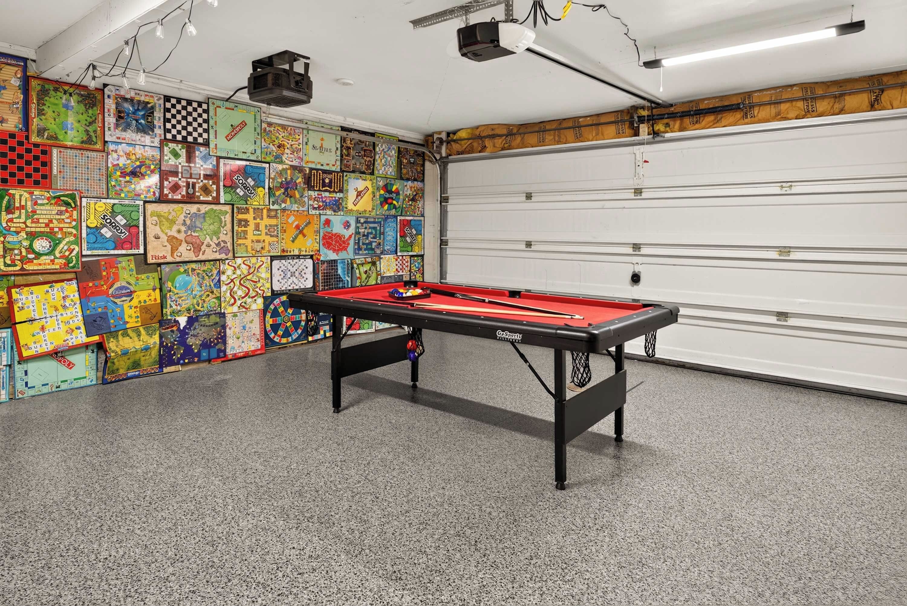A room with a pool table, a variety of board games on the wall, and a garage door.