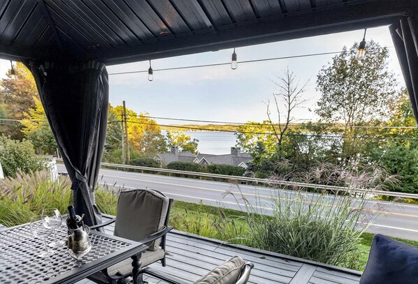 A covered outdoor seating area with a table, chairs, and a view of a road and trees. - Canandaigua Lake Overlook (Canandaigua)
