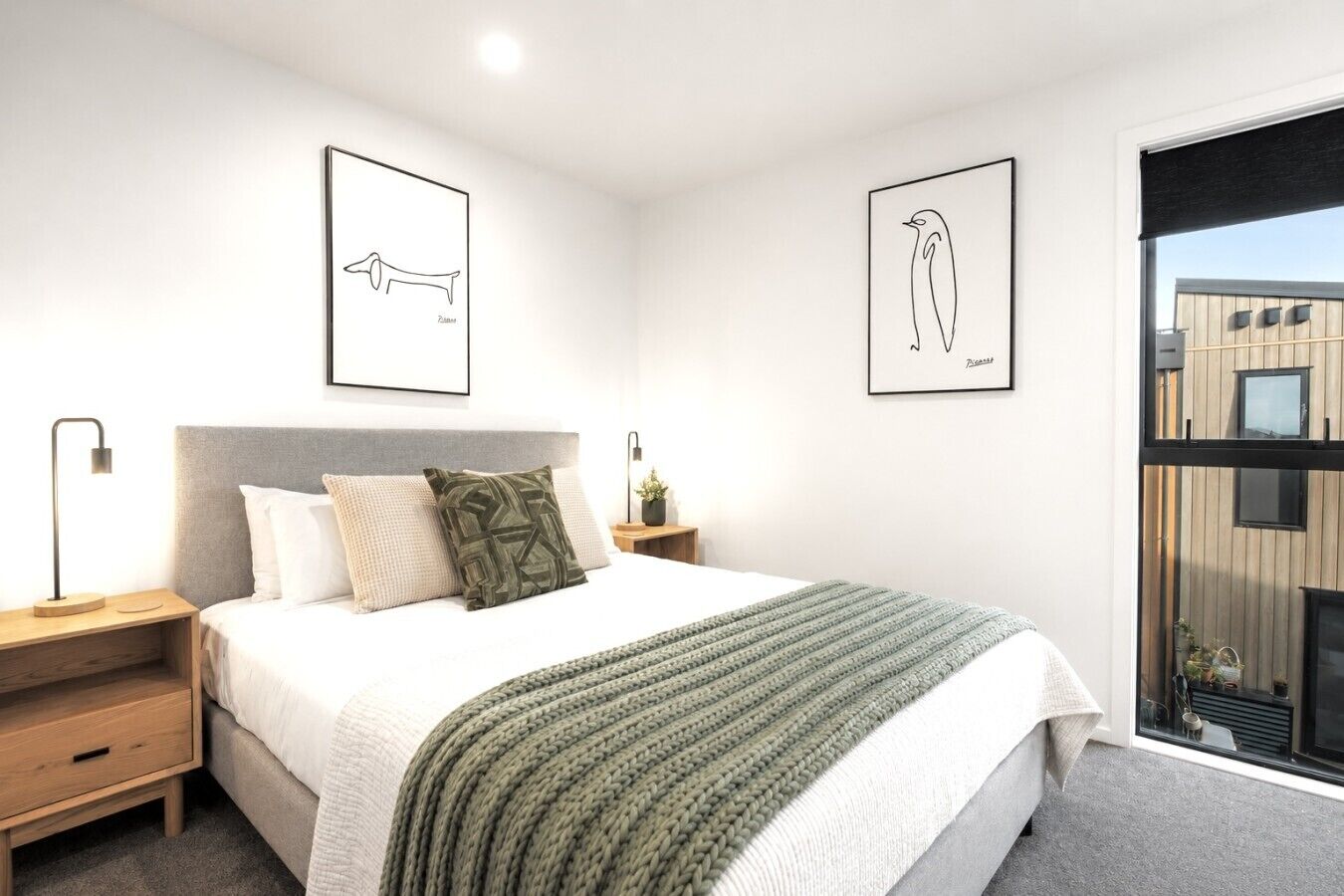 Une chambre à coucher moderne avec un lit, des tables de chevet, une lampe, une couverture verte en tricot et deux œuvres d’art encadrées accrochées au mur.