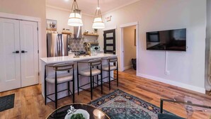 A modern kitchen with a central island, bar stools, stainless steel appliances, and a flat-screen TV mounted on the wall.