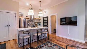 A modern kitchen with a central island, bar stools, stainless steel appliances, and a flat-screen TV mounted on the wall.