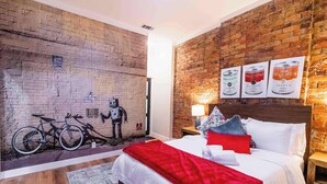 A hotel room with a brick wall mural, a bed with white and red bedding, a wooden floor, and a nightstand with a lamp.