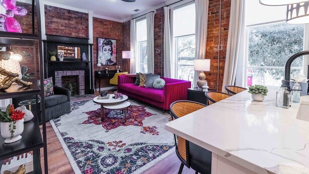 A modern living room with a brick wall, a fireplace, a vibrant pink sofa, and a large window with curtains.