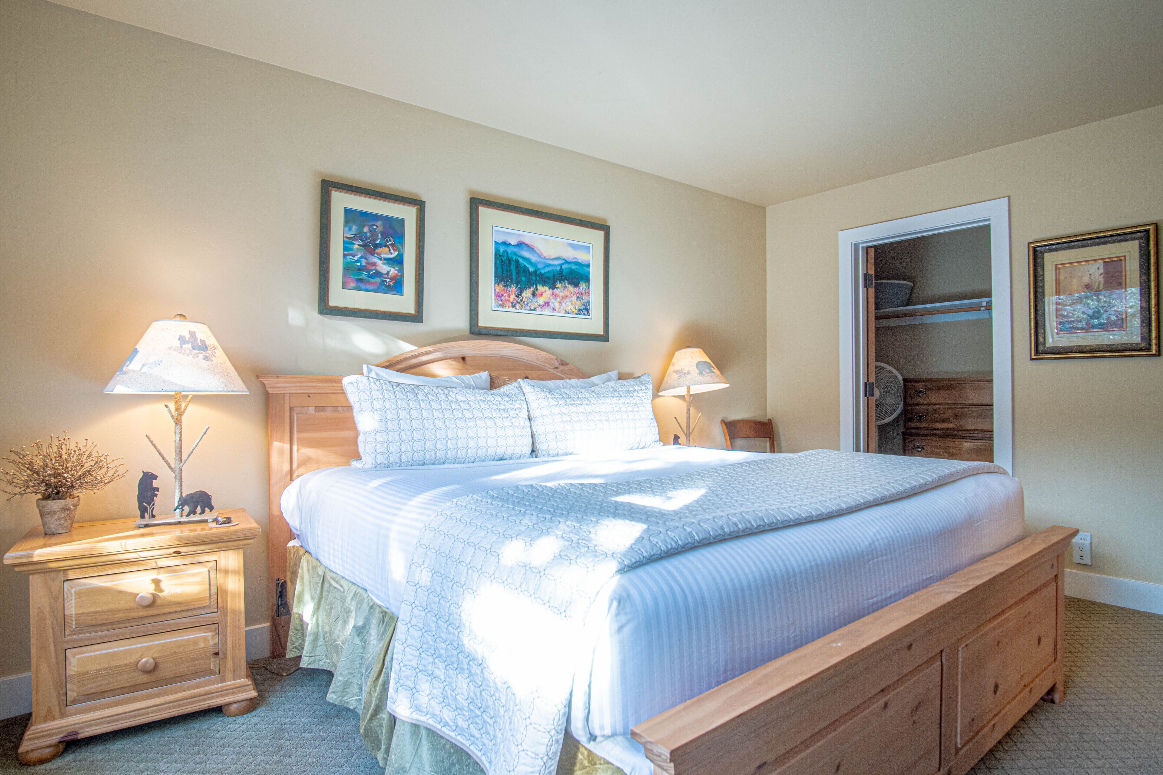 A bedroom with a wooden bed, two bedside tables with lamps, a nightstand with a plant, and two framed pictures on the wall.