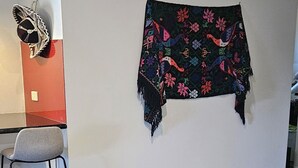 A dining area with a glass-top table, black chairs, and a colorful woven blanket on the wall.