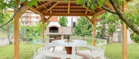 A covered outdoor dining area with a circular table and a brick oven in the background.