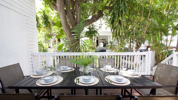 A dining table set for six with white plates, glasses, and cutlery, surrounded by wicker chairs, under a white fence with greenery in the background. - 3-bedroom townhome in beautiful Key West with AC and charm (Key West)