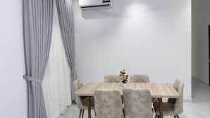 Un comedor con una mesa y sillas de color claro, un equipo de aire acondicionado en la pared y una cortina a la izquierda.