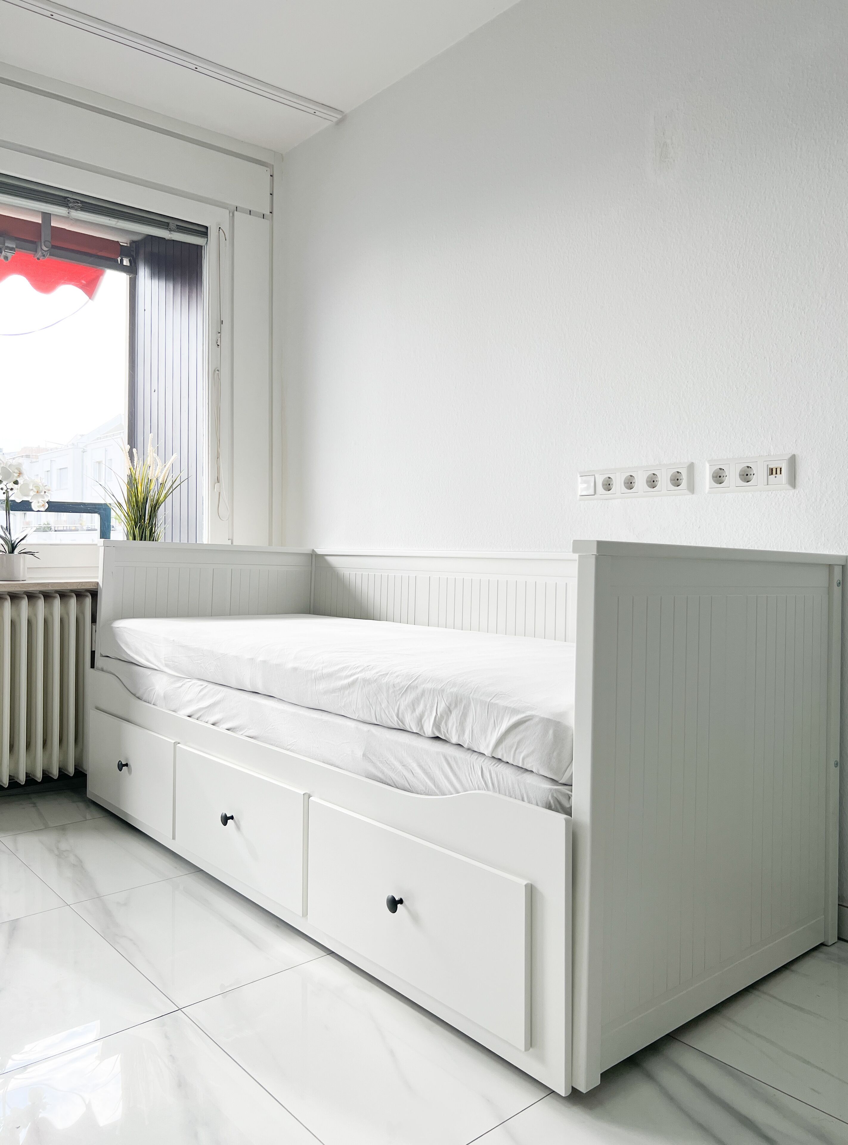 Un letto bianco con cassetti contenitori, un termosifone e una finestra con tenda rossa.