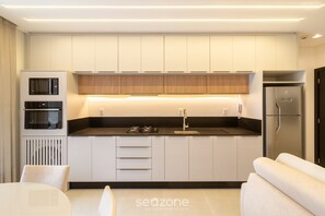 A modern kitchen with built-in appliances, a dining area, and a sofa. - Apto c\/ Varanda Gourmet próximo ao Mar OZN1004 (Itapema)