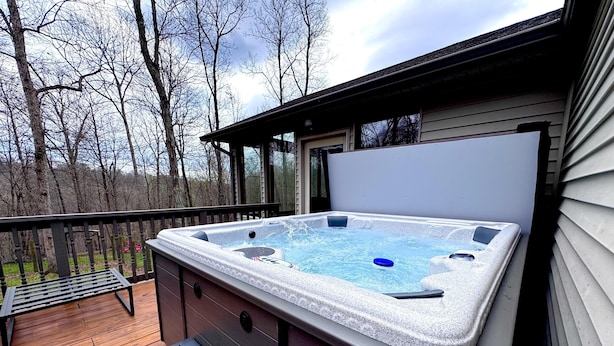 Een hottub op een houten terras met een bankje en een tafel, omgeven door bomen.