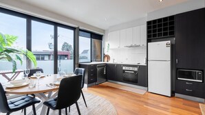 Een moderne keuken met zwarte kastjes, een ingebouwde oven en een koelkast. Er is een eethoek met een tafel en stoelen, en grote ramen bieden uitzicht op buiten.