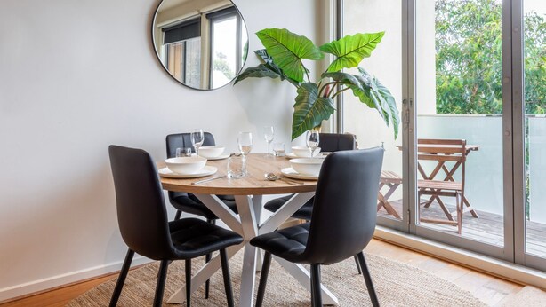 Een eetruimte met een ronde tafel, vier zwarte stoelen, witte borden en heldere glazen. Een grote potplant staat in de hoek, en er is een houten bank voor het raam.