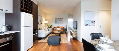 A modern apartment with a kitchen, dining area, and living room. It features wooden flooring, a sofa, a coffee table, and a TV.