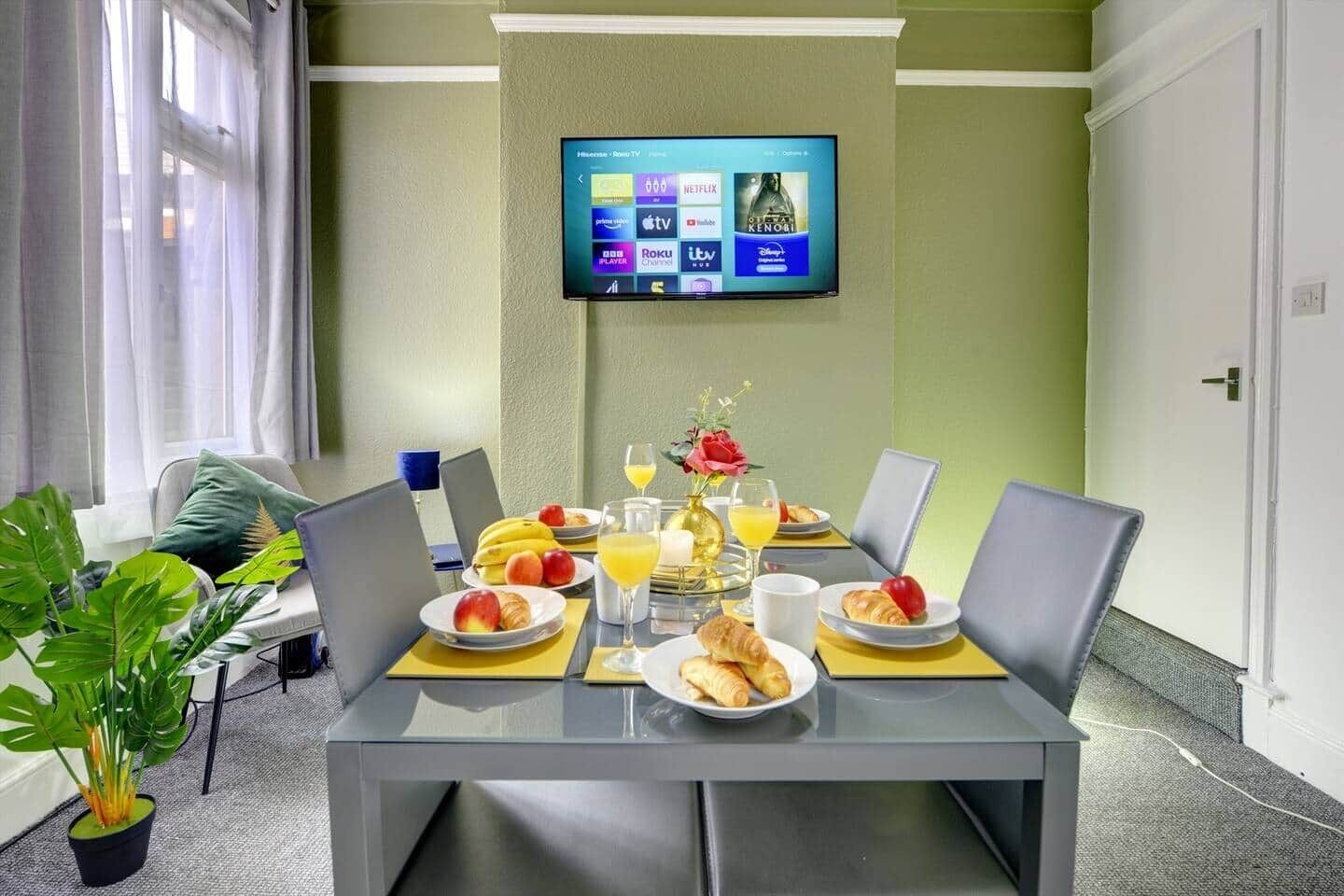 Une chambre d’hôtel avec une table à manger dressée pour le petit-déjeuner, un téléviseur à écran plat fixé au mur et une plante en pot posée sur le sol.