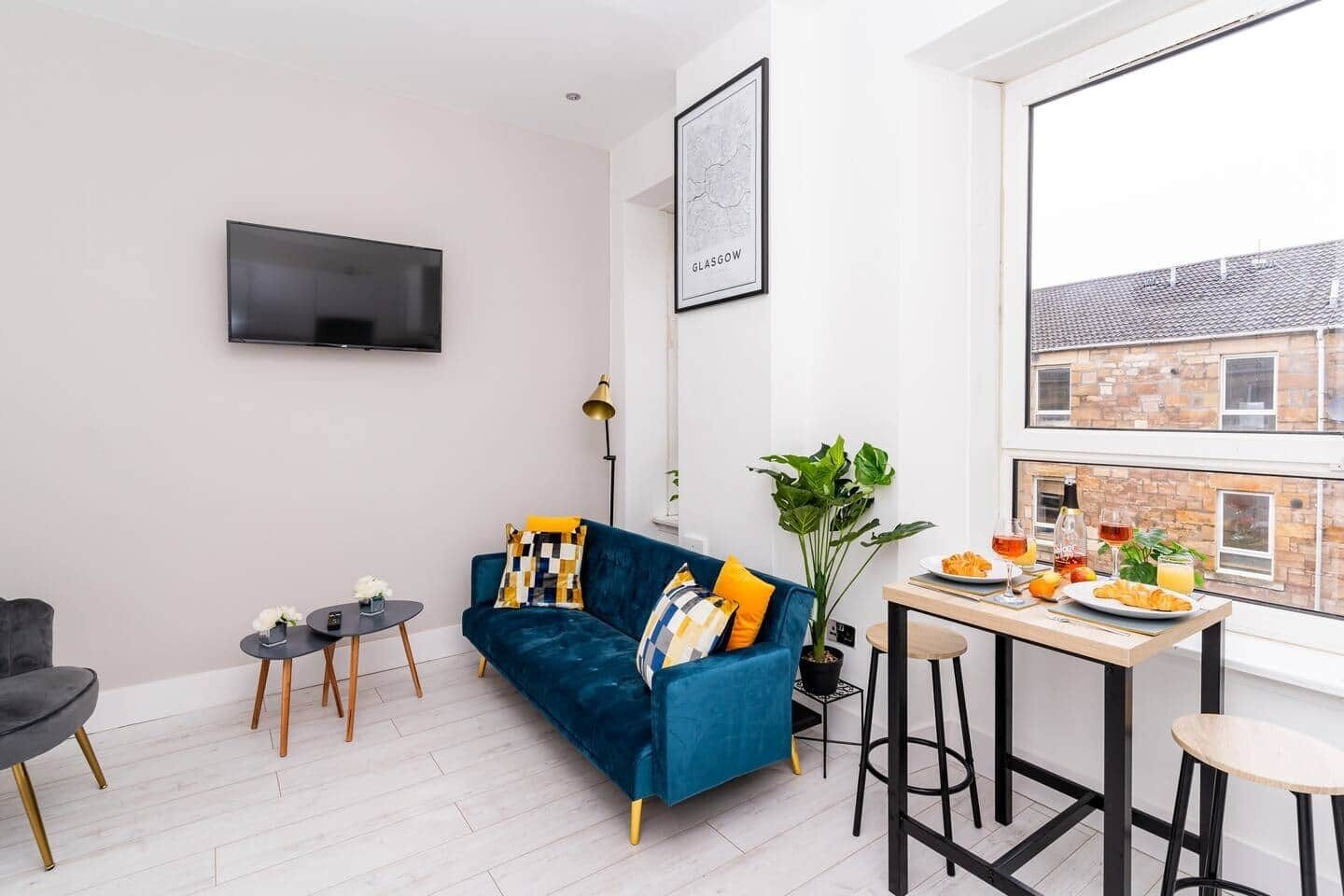 A modern living room with a blue sofa, a flat-screen TV, a dining table with breakfast items, and a large window with a city view.