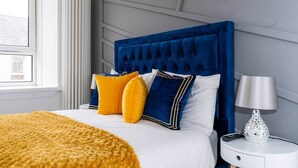 Un letto rifatto con cura, con una testiera blu, biancheria da letto bianca e cuscini gialli e blu. È presente un comodino bianco con una lampada e un telecomando.