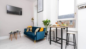 Un soggiorno moderno con un divano blu, un tavolo da pranzo con degli sgabelli, una pianta in vaso e vista sulla città attraverso una grande finestra.