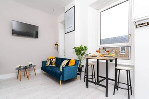 A modern living room with a blue sofa, a dining table with stools, a potted plant, and a cityscape view through a large window. - Arthur 2 Bed Home |Sleeps 4 Guest|Parking|3 Beds|Glasgow (Glasgow)