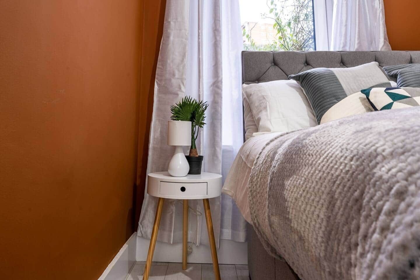 A bedroom with a bed, a bedside table with a lamp and a plant, and a window with curtains.