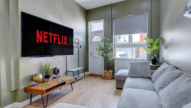 Een woonkamer met een grote flatscreen televisie aan de muur, een grijze bank, een houten salontafel en een potplant.
