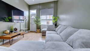 Een moderne woonkamer met een grijze bank, een houten salontafel, een aan de muur bevestigde televisie en twee potplanten.