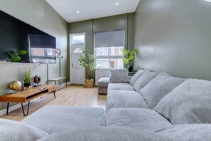 Ein modernes Wohnzimmer mit einer grauen Couch, einem Holztisch, einem an der Wand befestigten Fernseher und zwei Blumentöpfen.