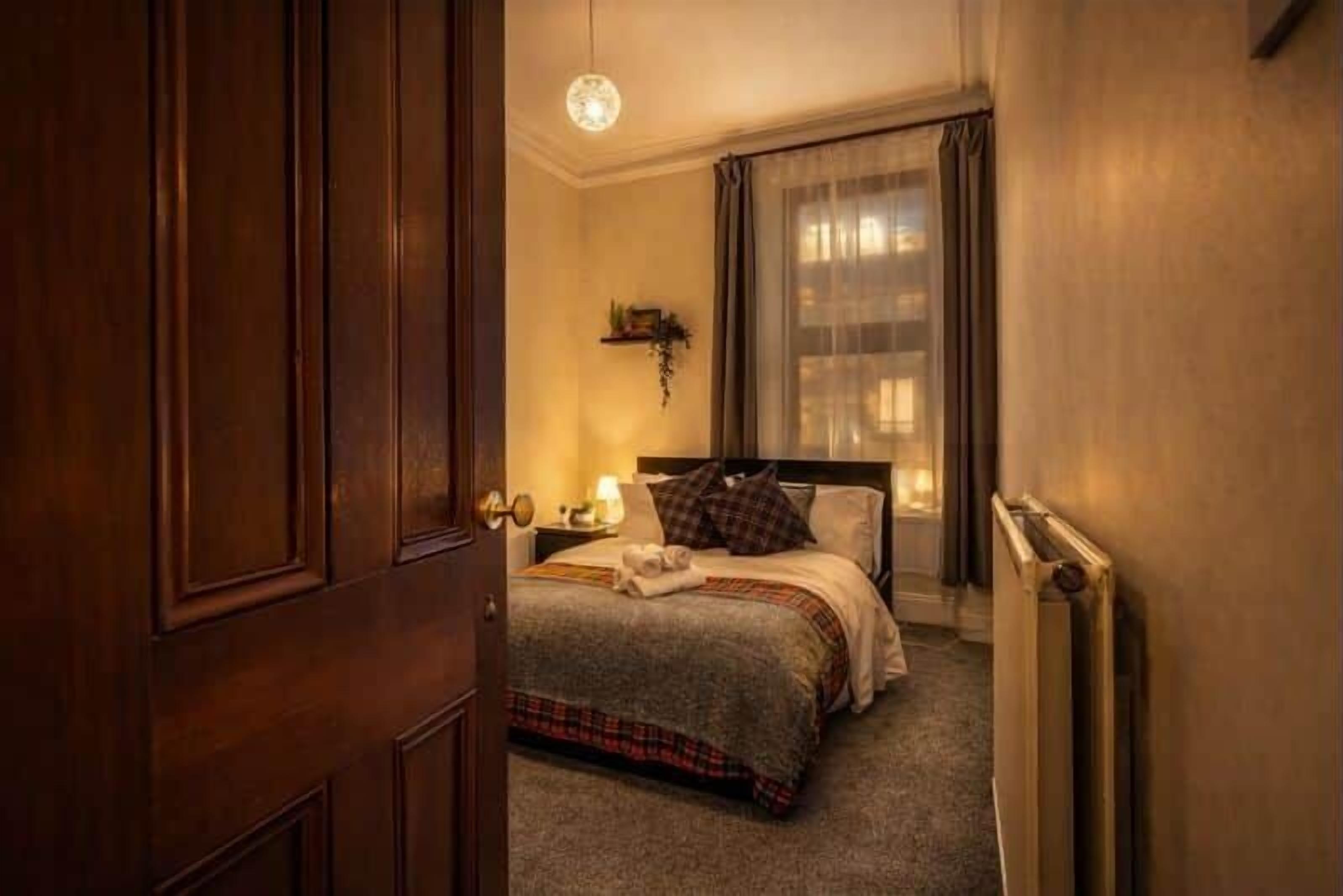 Una habitación de hotel con puerta de madera, cama con cubrecama a cuadros, mesita de noche con lámpara y ventana con cortinas traslúcidas.