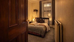 Quarto de hotel com porta de madeira, cama com colcha xadrez, mesa de cabeceira com abajur e janela com cortinas translúcidas.